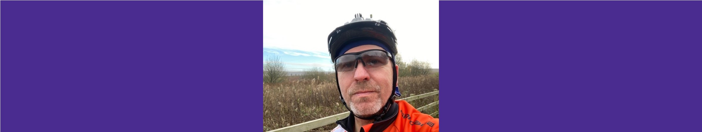Photo of Kevin wearing cycling gear, sunglasses and a helmet out in the countryside.